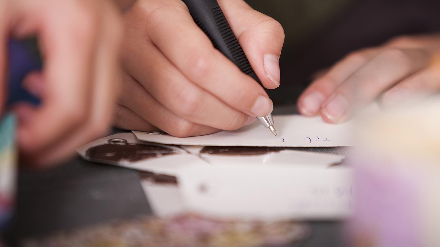 Nahaufnahme einer Hand, die mit einem Kugelschreiber auf einem Zettel schreibt, im Hintergrund eine brennende Kerze.